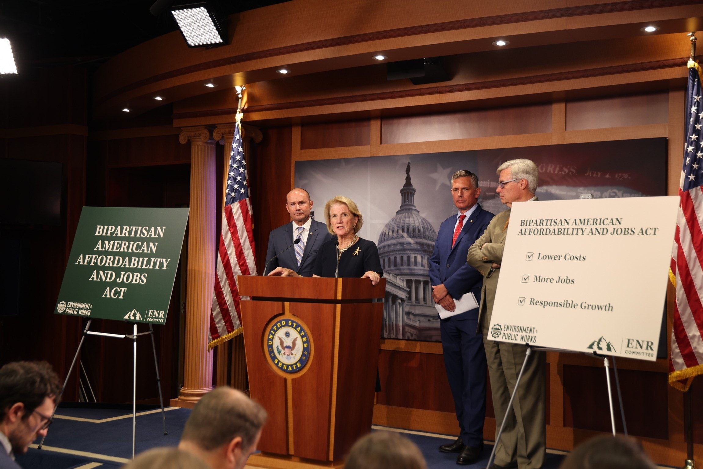 Speaker at a U.S. Senate podium during a press conference on the Bipartisan American Affordability and Jobs Act, with three people standing nearby, American flags, and signs highlighting lower costs, more jobs, and responsible growth.